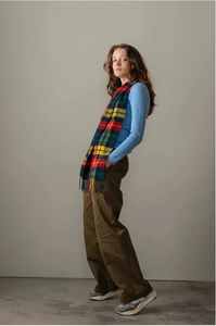 Person wearing a colorful plaid scarf, blue sweater, and brown pants against a plain background