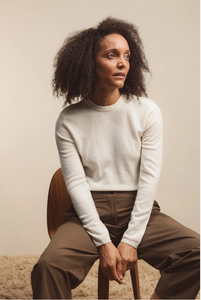 Woman sitting on a chair wearing a white long-sleeve top and brown pants against a beige background