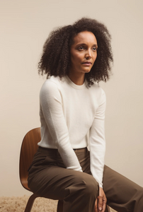 Woman sitting on a chair wearing a white sweater and brown pants against a plain background