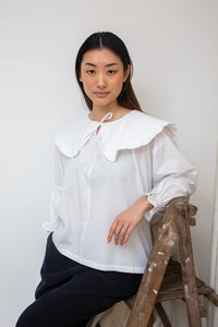Woman wearing a white blouse with a large collar sitting on a wooden ladder against a white wall.